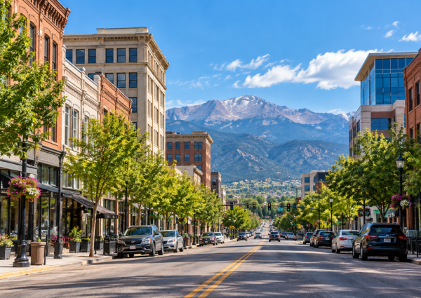 Tree-lined downtown Colorado Springs street with storefronts, office buildings, and mountain views in the distance.